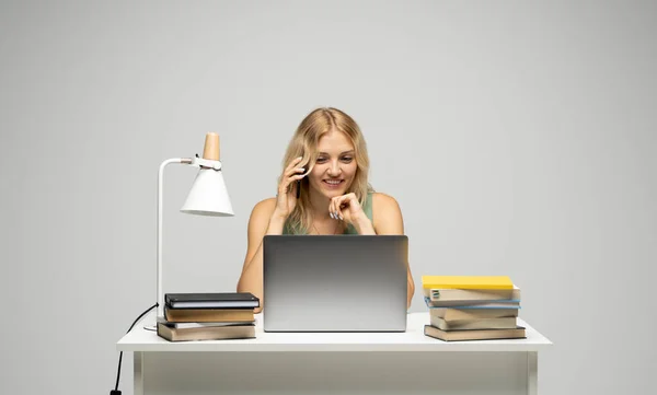 Joven mujer de negocios atractiva con un pelo rubio sentado en la mesa y trabajando en un ordenador portátil y hablando con un cliente en el teléfono. Joven estudiante alegre hablando con un teléfono inteligente. Estudiar. — Foto de Stock