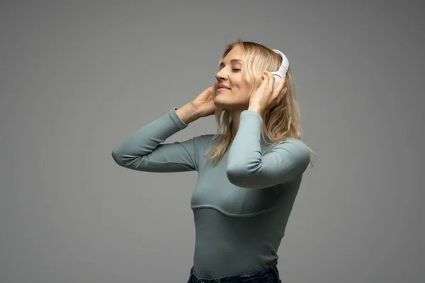 Hermosa mujer rubia joven atractiva con camiseta azul y gafas en auriculares blancos escuchando música y sonriendo sobre fondo gris en el estudio. Relajarse y disfrutar. Estilo de vida. —  Fotos de Stock