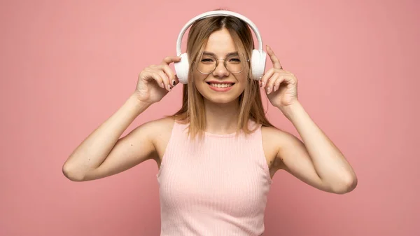 Hermosa mujer rubia joven atractiva con camiseta rosa y gafas en auriculares blancos escuchando música y sonriendo sobre fondo rosa en el estudio. Relajarse y disfrutar. Estilo de vida. —  Fotos de Stock