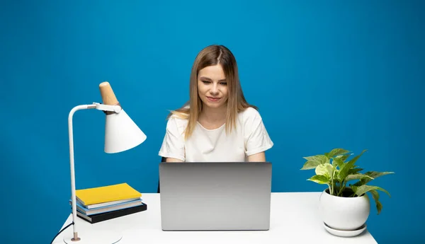 Retrato de uma jovem bonita estudando enquanto se senta na mesa com computador portátil cinza, notebook. Sorrindo mulher de negócios trabalhando com um laptop isolado em um fundo azul. — Fotografia de Stock
