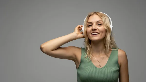 Belle jolie jeune femme blonde portant un t-shirt vert et des lunettes en casque blanc écoutant de la musique et souriant sur fond gris en studio. Détente et plaisir. Mode de vie. — Photo