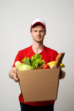 Kırmızı stüdyo arka planında, dükkandan alınmış yiyecek ve içeceklerle dolu bir paket taşıyan genç teslimatçı. Uzayı kopyala Teslimat Konsepti.