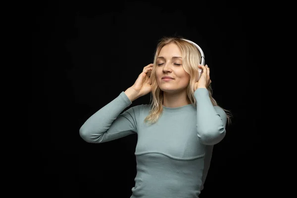 Hermosa mujer rubia joven atractiva con camiseta azul y gafas en auriculares blancos escuchando música y sonriendo sobre fondo negro en el estudio. Relajarse y disfrutar. Estilo de vida. —  Fotos de Stock
