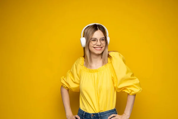 Krásná atraktivní mladá blondýnka ve žlutém tričku a brýlích v bílých sluchátkách, poslouchající hudbu, tančící a smějící se na modrém pozadí ve studiu. Uvolňující a užívající si. Životní styl. — Stock fotografie