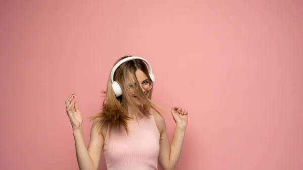 Krásná atraktivní mladá blondýna nosí růžové tričko a brýle v bílých sluchátkách, poslouchá hudbu a tančí na růžovém pozadí ve studiu. Uvolňující a užívající si. Životní styl. — Stock fotografie