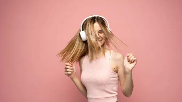 Hermosa mujer rubia joven atractiva con camiseta rosa y gafas en auriculares blancos escuchando música y bailando sobre fondo rosa en el estudio. Relajarse y disfrutar. Estilo de vida. —  Fotos de Stock
