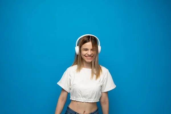 Jolie jeune femme blonde souriante portant un t-shirt blanc et des lunettes dans un casque blanc écoutant de la musique et dansant sur fond bleu en studio. Détente et plaisir. Mode de vie. — Photo