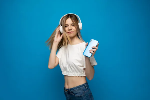 Hermosa mujer rubia joven atractiva con camiseta blanca y gafas en auriculares blancos escuchando música desde el teléfono inteligente, bailando y riendo sobre fondo azul en el estudio. Estilo de vida. —  Fotos de Stock