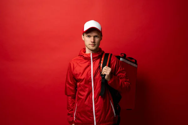 Fiatal futár, piros egyenruhás pólóban, piros kajás termo táskával a vállán, elszigetelve a vörös háttérstúdióban. Élelmiszer-szállítási szolgáltatás. — Stock Fotó