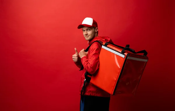 Dostawca facet pracownik w czerwonej czapce i jednolitej pracy odzieży roboczej jako kurier dealerski trzymać czerwoną torbę termiczną żywności i pokazuje kciuk w górę odizolowany na czerwonym tle studio kolor. Koncepcja usługi. — Zdjęcie stockowe