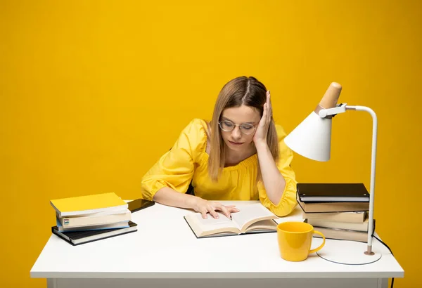 Уставшая молодая студентка в желтой повседневной одежде сидит за столом и читает книгу в библиотеке университета или колледжа. Сидя и читая на желтом фоне. Учёба. — стоковое фото