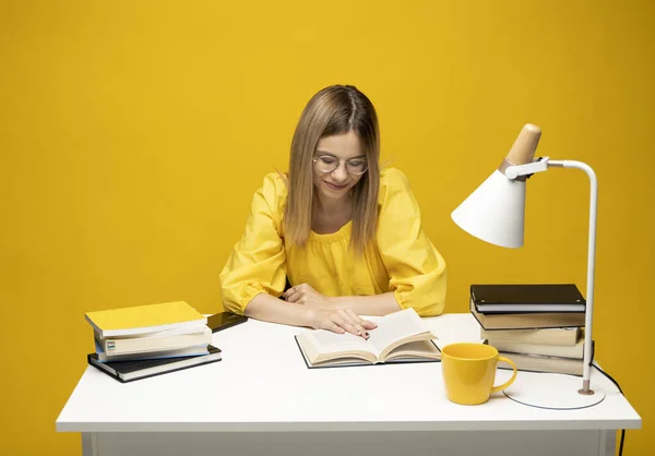 Молодая счастливая улыбающаяся студентка в желтой повседневной одежде читает книгу за столом в библиотеке университета или колледжа. Учёба. — стоковое фото