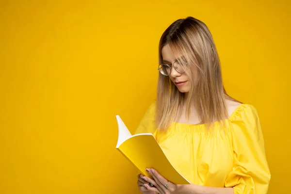 Портрет молодой студентки в жёлтой футболке и очках, читающей книгу на жёлтом фоне. Девушка читает роман. Учёба. — стоковое фото