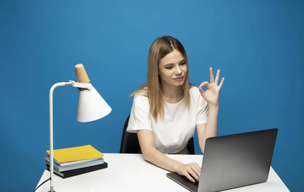 Retrato de uma empresária sorridente em uma camiseta branca trabalhando com um computador portátil e mostrando ok para colegas ou cliente enquanto fala com eles isolado sobre fundo azul. — Fotografia de Stock