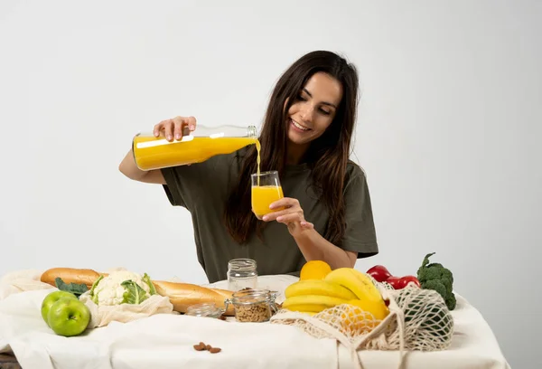 Mujer en camiseta verde vertiendo un jugo de una botella de vidrio en un vaso sobre una mesa con bolsa ecológica de malla, verduras veganas saludables, frutas, pan, bocadillos. Concepto de cero residuos. — Foto de Stock