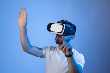 Siyah tişörtlü, 3D kulaklık takan bir adamın portresi. Sanal gerçeklikte video oyunları oynarken yaptığı bir şeye dokunur gibi el kol hareketi yapıyor. Metaevren.
