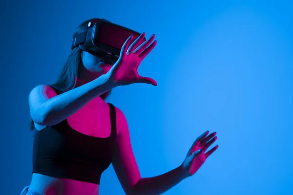 Młoda kobieta w wirtualnej rzeczywistości w neonowym świetle na niebieskim tle. — Zdjęcie stockowe