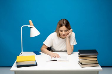 Üniversite kütüphanesinde ya da üniversitede, beyaz günlük giysiler içinde kitap okuyan genç bir bayan öğrenci. Mavi arka planda oturuyor ve okuyor. Çalışıyorum.