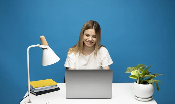Podekscytowana młoda kobieta pracująca z szarym laptopem, notatnik siedząc przy stole z szarym laptopem. Uśmiechnięta kobieta biznesowa lub student otrzymał dobrą wiadomość izolowane na niebieskim tle. — Zdjęcie stockowe