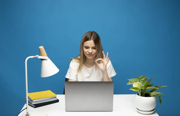 Está tudo bem. Mulher feliz com uma camisa branca e óculos mostrando sinal ok, sentado com laptop sobre fundo azul. — Fotografia de Stock