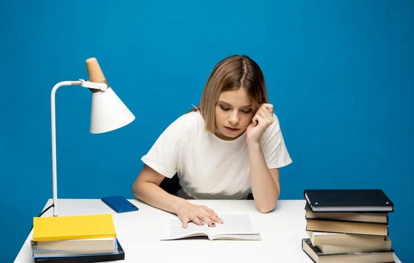 Уставшая молодая студентка в белой повседневной одежде сидит за столом и читает книгу в библиотеке университета или колледжа. Сидя и читая на синем фоне. Учёба. — стоковое фото
