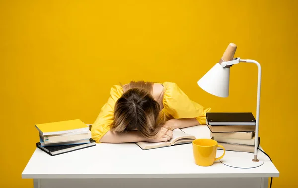 Студент, изучающий экзамен и спящий на книгах на желтом фоне. Уставшая студентка в очках спит на книгах в библиотеке. — стоковое фото