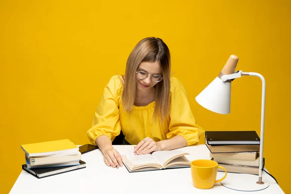 Junge glücklich lächelnde Studentin in gelber Freizeitkleidung, die das Buch am Tisch in der Bibliothek der Universität oder Hochschule liest. Studieren. — Stockfoto