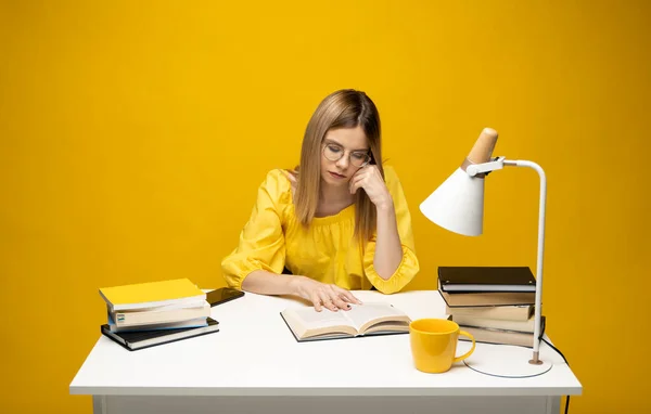 Уставшая молодая студентка в желтой повседневной одежде сидит за столом и читает книгу в библиотеке университета или колледжа. Сидя и читая на желтом фоне. Учёба. — стоковое фото