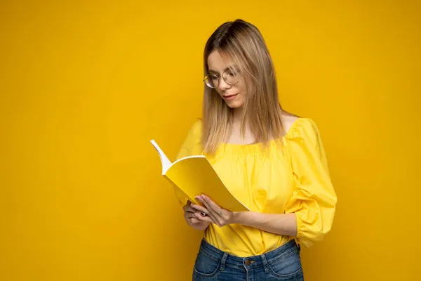 Сосредоточенная молодая женщина-подросток в очках читает интересную книгу, изолированную на жёлтом фоне. Девушка читает роман. Учёба. — стоковое фото