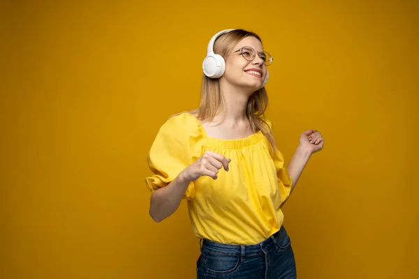 Jovencita alegre bonita sonriendo mientras escucha música en auriculares y bailando sobre fondo amarillo. —  Fotos de Stock