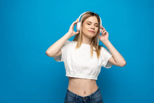 Hermosa mujer joven en auriculares escuchando música de fondo azul. Joven hermosa mujer en traje brillante disfrutando de una música. —  Fotos de Stock