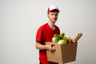 Teslimat Konsepti. Kırmızı üniformalı memnun teslimatçının portresi beyaz arka planda izole edilmiş yiyecek ürünleriyle dolu kağıt poşet taşırken gülümsüyor. Yemek dağıtım servisi..