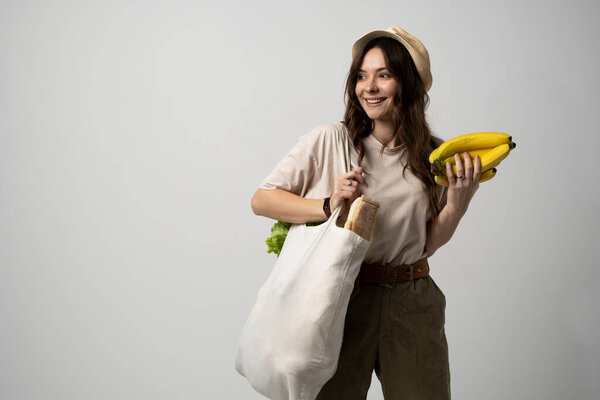 Zero waste concept. Young brunette woman holding cotton eco bag with organic fruits and vegetables on a shoulder and holding bananas in a hand. Using reusable crochet net bag for grocery shopping.