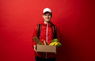 Kırmızı üniformalı teslimatçı, beyaz arka planda karton karton karton kutu ve stüdyo portresi tutuyor. Servis kavramı.