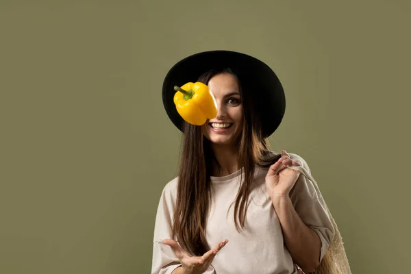 Eco friendly smiling woman in beige t-shirt and black hat throw up a yellow pepper in a air by one hand and holding reusable cotton eco bags full of groceries on a shoulder. — Stock Photo, Image