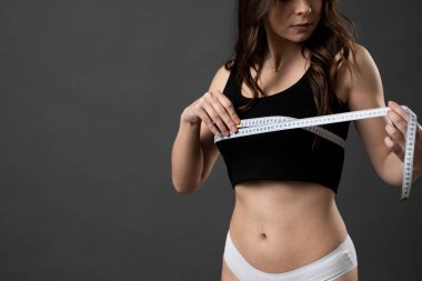 Siyah bluzlu ve beyaz külotlu genç esmer kadın göğüslerini mezurayla ölçüyor..