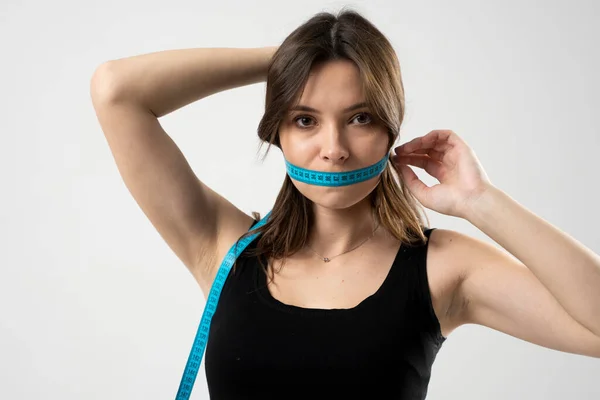 Woman in a black top and panties with a blue measuring tape over a mouth, in a white background. Healthy lifestyle concept. Diet.
