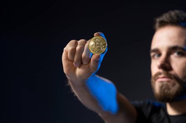 Siyah tişört giyen, elinde altın Bitcoin tutan ve siyah arka planda tek başına duran olgun sakallı bir adam..