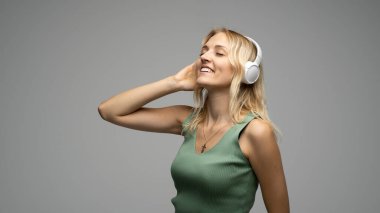 Beyaz kulaklıklı sarışın kız stüdyodaki gri arka planda kapalı gözlerle müzik dinliyor. Gri tişört giyiyor..