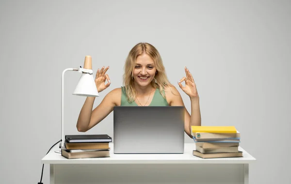 Estudiante mostrando gesto bien con ambas manos, feliz con el trabajo terminado, sonriendo y mirando a la cámara mientras trabaja en el ordenador portátil sentado frente a grandes pilas de libros. — Foto de Stock