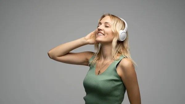 Blondes Mädchen mit weißen Kopfhörern, das mit geschlossenen Augen im Studio Musik auf grauem Hintergrund hört. Sie trägt ein graues T-Shirt. — Stockfoto