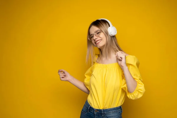 Mujer rubia con gafas y camisa amarilla escuchando música con auriculares. Adolescente chica bailando sobre amarillo fondo. —  Fotos de Stock