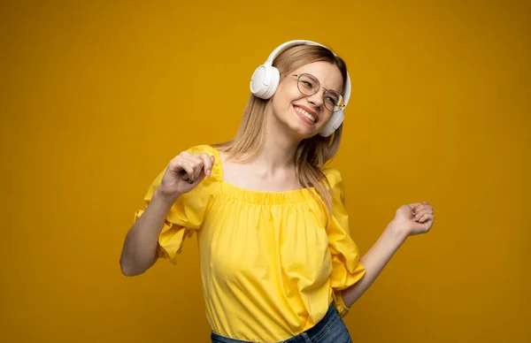 Jovencita alegre bonita sonriendo mientras escucha música en auriculares y bailando sobre fondo amarillo. —  Fotos de Stock