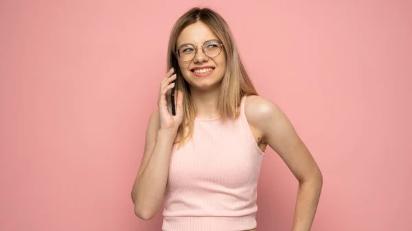 Photo rapprochée d'une jolie jeune femme blonde riante et positive portant une chemise jaune élégante et des lunettes communiquant sur un téléphone portable et souriant isolé sur fond jaune. — Photo