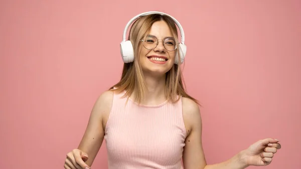 Menina bonita em uma camiseta rosa e óculos ouvindo música com seus fones de ouvido e dançando em fundo rosa. — Fotografia de Stock