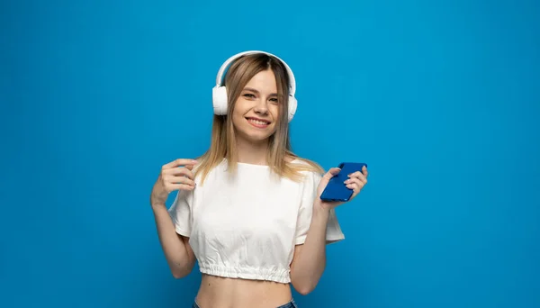 Mosolygó lány pihentető otthon, ő játszik zenét használ okostelefon és fehér vezeték nélküli fejhallgató. — Stock Fotó
