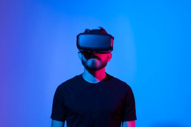 Meta-evren ve yaşamsal gerçeklik kavramı. VR gözlüklü Avrupalı sakallı adam, sanal gerçeklik kulaklığıyla video oyunları oynuyor, pembe mavi renkli bir şeye dokunmaya çalışıyor..