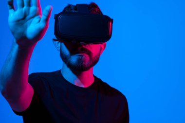 Artırılmış gerçeklik ve sanal gerçeklik dünyası. VR gözlüklü heyecanlı sakallı adam, gözlüklü video oyunu oynarken sanal düğmeye basıyor, mor arka planda neon ışığı altında duruyor..