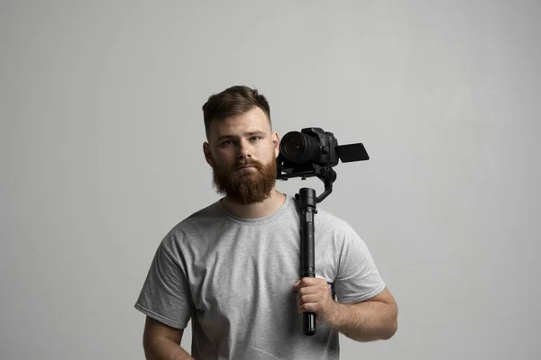 Ritratto di cineasta barbuto in t-shitr grigio con fotocamera e gimbal a 3 assi su sfondo bianco. Videografia, Filmmaking, hobby e creatività. — Foto Stock