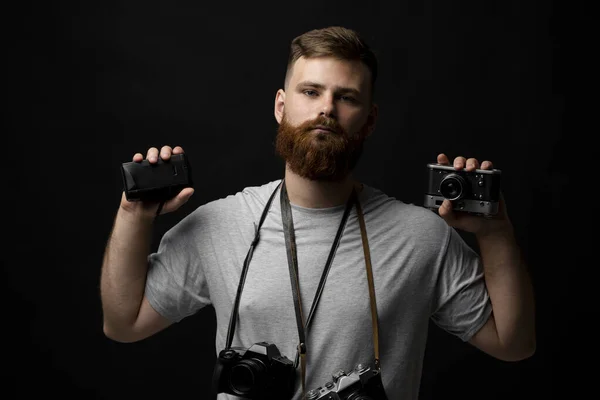Professional photographer in a grey t-shirt with a bunch of different cameras in a hands and on a shoulder looking on a camera and ready for make a good shoot on a black background. — Stock Photo, Image